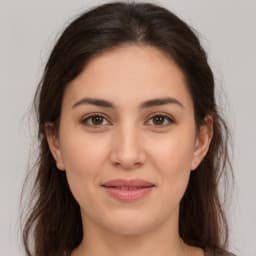 Bu resim koyu renk saçlı genç bir kadının yakın plan portresidir. Oval bir yüz şekli var ve gözleri kalın kaşlarla çerçevelenmiş. Dudakları nazik bir gülümsemeyle hafifçe ayrılmış, beyaz dişleri ve pembe ruju ortaya çıkıyor. Yanaklarında pembe bir ton var ve cildi pürüzsüz ve kusursuz görünüyor. Işık, yumuşak dalgalar halinde omuzlarına dökülen saçlarındaki ışıltıları yakalıyor. Doğal güzelliğini aşırıya kaçmadan ortaya çıkaran minimal bir makyaj yapıyor. Gömleğinin yakasının altından görünen boynu, üzerinde küçük takılar bulunan narin bir kolye taktığı göğsünün üst kısmına kadar iniyor. Bu portre, gençliğin ve neşenin özünü yakalıyor, çünkü bu kadın kamera objektifine kendinden emin bir şekilde gülümsüyor - kendinden emin ama aynı zamanda savunmasız - zaman içinde sonsuza dek yakalanan unutulmaz bir an yaratıyor.